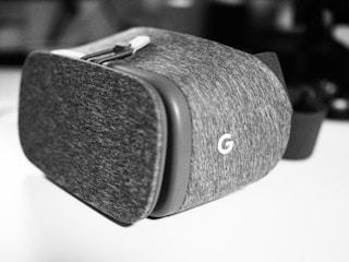 A close-up view of a virtual reality headset with a fabric exterior and an embossed 'G' logo. The headset has a strap attached, indicating it is designed to be worn on the head.