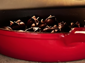 Roasted chestnuts are nestled in a red ceramic dish, with their shells cracked open, revealing an appetizing golden interior. The dish is set against a neutral background, highlighting the warm and rustic atmosphere.