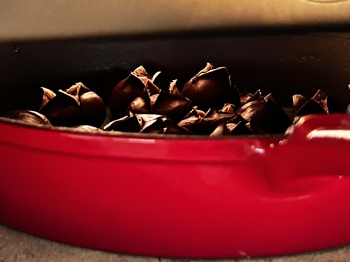 Roasted chestnuts are nestled in a red ceramic dish, with their shells cracked open, revealing an appetizing golden interior. The dish is set against a neutral background, highlighting the warm and rustic atmosphere.