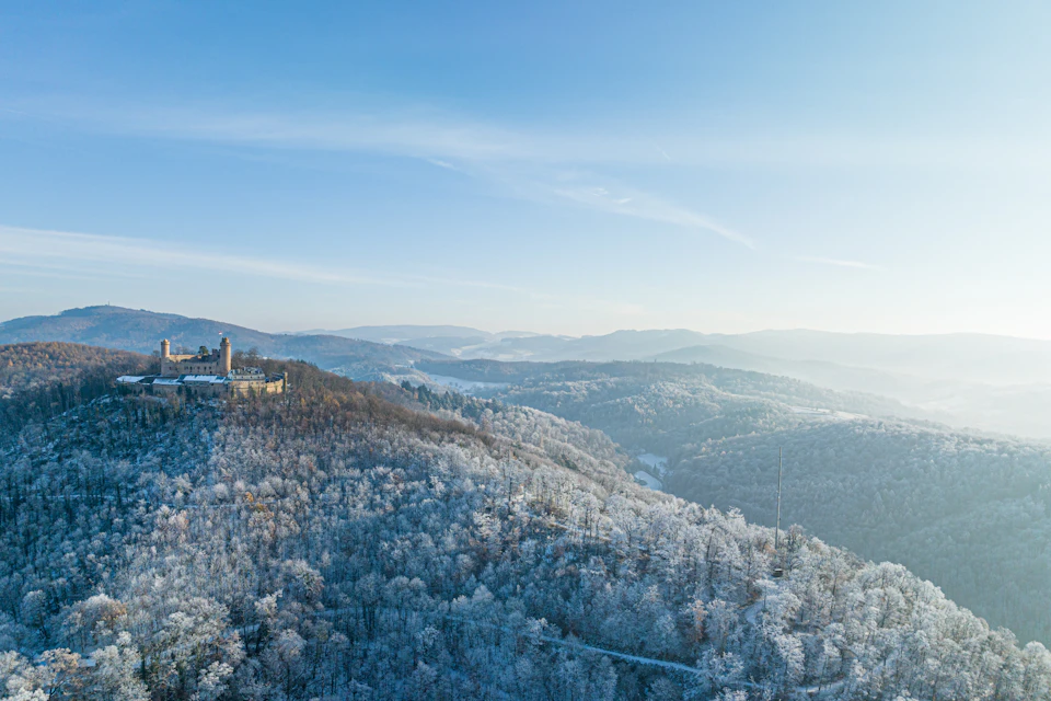 The image presents an aerial view of a mountainous landscape, with a castle-like structure nestled amidst the verdant trees and rolling hills. The castle is perched on a hilltop, surrounded by a dense forest that blankets the area in a thick layer of snow. The sky above mirrors the color of the snow below, creating a serene and tranquil atmosphere.