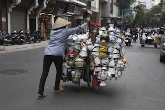 A person wearing a conical hat is pushing a bicycle loaded with various pottery and ceramic items on a street in an urban area. Shops and motorcycles are visible in the background, and other people are also using the street, creating a bustling atmosphere.