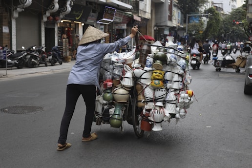 A person wearing a conical hat is pushing a bicycle loaded with various pottery and ceramic items on a street in an urban area. Shops and motorcycles are visible in the background, and other people are also using the street, creating a bustling atmosphere.