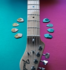 A guitar neck lies diagonally across a colorful background split into teal and magenta sections. Guitar picks in various shades of blue and beige are arranged on both sides of the neck.