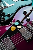 A close-up view of two electric guitars, one black and the other a glossy purple. The purple guitar has three colorful picks placed on it—yellow, orange, and turquoise. The black guitar features multiple control knobs and a metallic bridge, with its polished surface reflecting light.