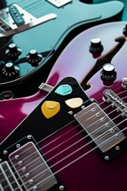 A close-up view of two electric guitars, one black and the other a glossy purple. The purple guitar has three colorful picks placed on it—yellow, orange, and turquoise. The black guitar features multiple control knobs and a metallic bridge, with its polished surface reflecting light.