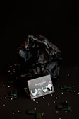 A dark, moody composition features a package labeled 'ROMBO TRY OUT MIX ECO-BLACK' with four guitar picks displayed. The background includes a crumpled black material, with small transparent beads and dark green rectangular pieces scattered around, adding texture and depth.