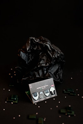 A dark, moody composition features a package labeled 'ROMBO TRY OUT MIX ECO-BLACK' with four guitar picks displayed. The background includes a crumpled black material, with small transparent beads and dark green rectangular pieces scattered around, adding texture and depth.