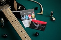 A black electric guitar is placed on a green surface, accompanied by a guitar cable with a gold plug, two cards with graphics and text about guitar picks, and electronic components like vacuum tubes and knobs. The scene is well-organized and artistically composed, highlighting musical equipment.
