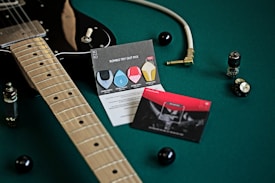 A black electric guitar is placed on a green surface, accompanied by a guitar cable with a gold plug, two cards with graphics and text about guitar picks, and electronic components like vacuum tubes and knobs. The scene is well-organized and artistically composed, highlighting musical equipment.