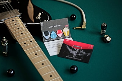 A black electric guitar is placed on a green surface, accompanied by a guitar cable with a gold plug, two cards with graphics and text about guitar picks, and electronic components like vacuum tubes and knobs. The scene is well-organized and artistically composed, highlighting musical equipment.