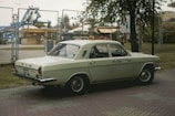 A vintage, cream-colored car with a taxi sign on top is parked near a fenced area. The background includes an amusement park with visible rides and structures, and a tree partially covers the view on the right.