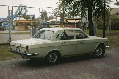 A vintage, cream-colored car with a taxi sign on top is parked near a fenced area. The background includes an amusement park with visible rides and structures, and a tree partially covers the view on the right.