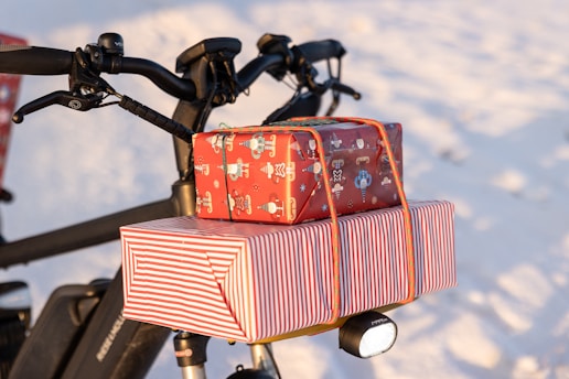 A bicycle with two wrapped gifts secured on the rear rack. The larger gift is wrapped in red and white striped paper, while the smaller one features festive holiday motifs on red paper. Snow covers the ground, suggesting a winter setting.