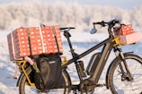 A bicycle loaded with several gift-wrapped packages against a snowy background. The bicycle features a black frame and panniers, with large red and white presents secured on the back and front. There is snow on the ground, and trees are visible in the distance, suggesting a winter setting.
