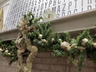 A festive Christmas garland decorates a white mantle, featuring green pine leaves, gold ribbon, pine cones, and ornaments. Above the mantle, a wall is covered with a decorative white board featuring large black letters arranged in a random pattern. A decorative star is placed above the garland.