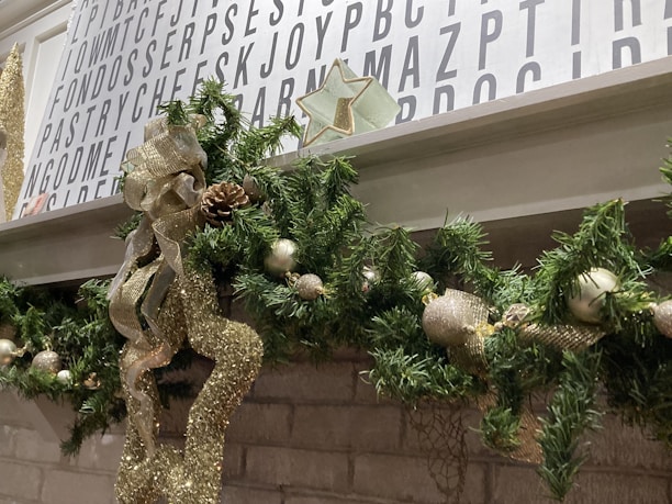 A festive Christmas garland decorates a white mantle, featuring green pine leaves, gold ribbon, pine cones, and ornaments. Above the mantle, a wall is covered with a decorative white board featuring large black letters arranged in a random pattern. A decorative star is placed above the garland.