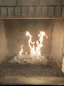 A fireplace with visible flames burning on a bed of dark, shiny stones inside a mesh enclosure. The background consists of brickwork, adding a rustic feel.