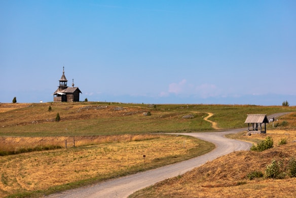 A rural landscape featuring a small wooden church with a steeple situated on a grassy hill. A winding dirt road leads toward the church, with another branching path leading to a wooden shelter. The terrain consists of dried grass with some patches of greenery under a clear, blue sky.