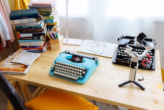 A wooden desk is cluttered with two typewriters, one modern and one vintage. A blue mechanical typewriter stands out. Stacks of books and colorful stationery fill the desk. A gimbal stabilizer with a smartphone is positioned in the right foreground, adding a tech touch. Sunlight filters through sheer curtains, creating a cozy workspace ambiance.