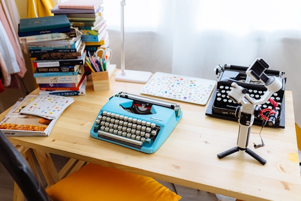 A wooden desk is cluttered with two typewriters, one modern and one vintage. A blue mechanical typewriter stands out. Stacks of books and colorful stationery fill the desk. A gimbal stabilizer with a smartphone is positioned in the right foreground, adding a tech touch. Sunlight filters through sheer curtains, creating a cozy workspace ambiance.