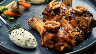 A gourmet dish featuring roasted chicken drumsticks topped with sesame seeds and walnuts, served with a dollop of creamy sauce sprinkled with poppy seeds on a dark plate. Garnishes include fresh rosemary and a side of sautéed vegetables, including carrots, mushrooms, and broccoli.