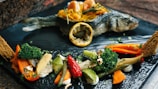 A whole cooked fish is elegantly presented on a black plate, accompanied by a variety of vegetables and garnished with a slice of lemon. The plate features colorful vegetables like broccoli, baby corn, red and orange bell peppers, green beans, and what appears to be gold leaf accents. Bread crisps are also visible alongside the dish.