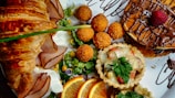 A delicious spread of brunch items features a flaky croissant with slices of ham, fresh salad greens with corn, mini quiches topped with melted cheese and garnish, crispy fried balls, and pancakes drizzled with chocolate and topped with a strawberry. Slices of orange add a fresh citrus touch.