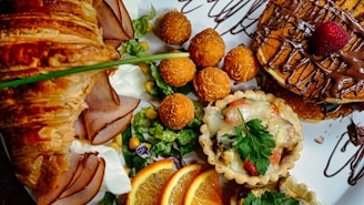 A delicious spread of brunch items features a flaky croissant with slices of ham, fresh salad greens with corn, mini quiches topped with melted cheese and garnish, crispy fried balls, and pancakes drizzled with chocolate and topped with a strawberry. Slices of orange add a fresh citrus touch.