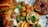 A delicious spread of brunch items features a flaky croissant with slices of ham, fresh salad greens with corn, mini quiches topped with melted cheese and garnish, crispy fried balls, and pancakes drizzled with chocolate and topped with a strawberry. Slices of orange add a fresh citrus touch.