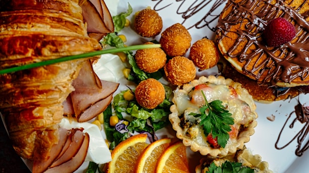 A delicious spread of brunch items features a flaky croissant with slices of ham, fresh salad greens with corn, mini quiches topped with melted cheese and garnish, crispy fried balls, and pancakes drizzled with chocolate and topped with a strawberry. Slices of orange add a fresh citrus touch.