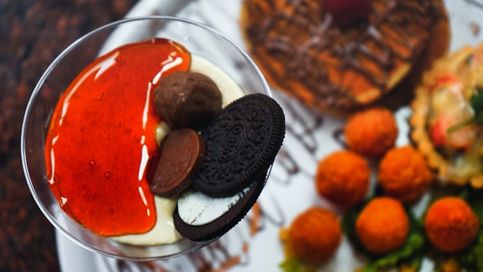 A dessert spread featuring a glass of creamy pudding topped with a vibrant red sauce, chocolate truffles, and a black sandwich cookie. Surrounding the glass are various other desserts, including a chocolate-drizzled pastry and some small orange-hued balls.