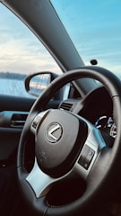 A close-up view of a car's interior focusing on the steering wheel with a Lexus logo. The dashboard is visible in the background, and a side mirror reflects the snowy landscape outside. The sky is clear with a few clouds, indicating a cold but sunny day.