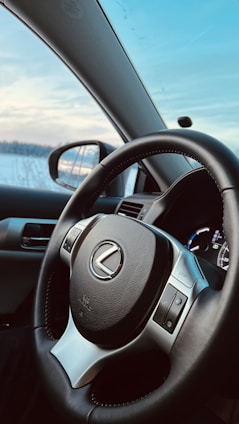 A close-up view of a car's interior focusing on the steering wheel with a Lexus logo. The dashboard is visible in the background, and a side mirror reflects the snowy landscape outside. The sky is clear with a few clouds, indicating a cold but sunny day.
