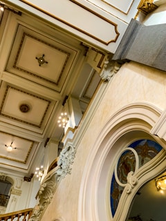 An intricately designed interior with ornate architectural details, including a high ceiling with decorative molding and light fixtures. A curved archway features stained glass and sculpted embellishments.