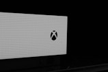 A close-up view of a white, perforated electronic device with a logo resembling a stylized 'X' on its front. The image is predominantly monochrome, with the device against a black background, emphasizing the contrast.