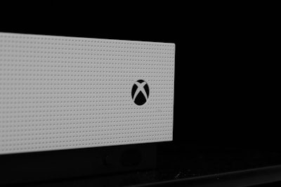 A close-up view of a white, perforated electronic device with a logo resembling a stylized 'X' on its front. The image is predominantly monochrome, with the device against a black background, emphasizing the contrast.