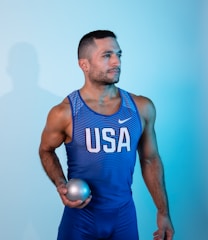 A man in athletic wear is holding a shot put ball. He is wearing a blue and white sports tank top with 'USA' printed on it and matching blue shorts. The background is a gradient of light blue, creating a cool-toned aesthetic.