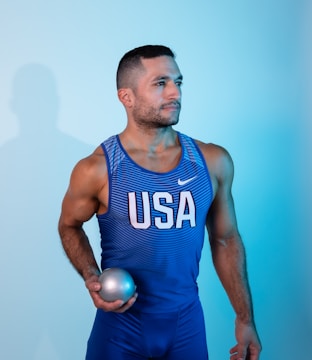 A man in athletic wear is holding a shot put ball. He is wearing a blue and white sports tank top with 'USA' printed on it and matching blue shorts. The background is a gradient of light blue, creating a cool-toned aesthetic.