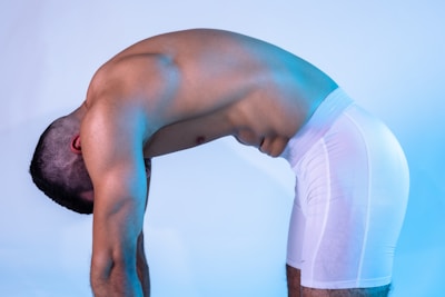 A muscular individual is bent forward, showing a flexed back and wearing white shorts. The background is a light blue, creating a calm atmosphere.