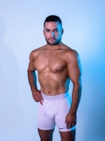 A muscular man stands confidently against a light blue background, wearing white athletic shorts. The lighting highlights his physique, casting soft shadows.