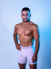 A muscular man stands confidently against a light blue background, wearing white athletic shorts. The lighting highlights his physique, casting soft shadows.