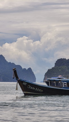 A wooden boat labeled 'Guide Nong' is floating on a calm body of water, surrounded by lush green hills and dramatic, cloud-filled skies. The scene appears tranquil and serene.