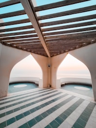 An architectural space with a pergola-like wooden structure overhead and white arched walls. The floor is tiled with alternating stripes of teal green and white tiles. Two large circular openings offer a view of a serene sky and horizon in the background.