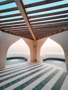 An architectural space with a pergola-like wooden structure overhead and white arched walls. The floor is tiled with alternating stripes of teal green and white tiles. Two large circular openings offer a view of a serene sky and horizon in the background.
