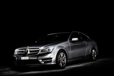 A silver luxury car is set against a dark background, highlighted dramatically by focused lighting that emphasizes its sleek and shiny bodywork.