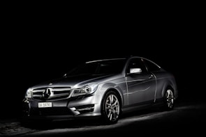 A silver luxury car is set against a dark background, highlighted dramatically by focused lighting that emphasizes its sleek and shiny bodywork.