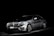 A silver luxury car is set against a dark background, highlighted dramatically by focused lighting that emphasizes its sleek and shiny bodywork.