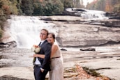 A couple dressed in formal wedding attire poses in front of a cascading waterfall amidst a natural rocky landscape. The bride holds a bouquet of flowers and leans affectionately against the groom, who is wearing a suit. The scene is set against a backdrop of lush greenery and flowing water, with autumn leaves scattered across the ground.