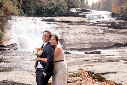 A couple dressed in formal wedding attire poses in front of a cascading waterfall amidst a natural rocky landscape. The bride holds a bouquet of flowers and leans affectionately against the groom, who is wearing a suit. The scene is set against a backdrop of lush greenery and flowing water, with autumn leaves scattered across the ground.