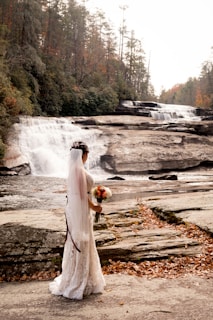 A bride stands on a rocky outcrop near a waterfall, surrounded by autumn foliage. She is wearing a lace wedding dress and veil, holding a bouquet of flowers.
